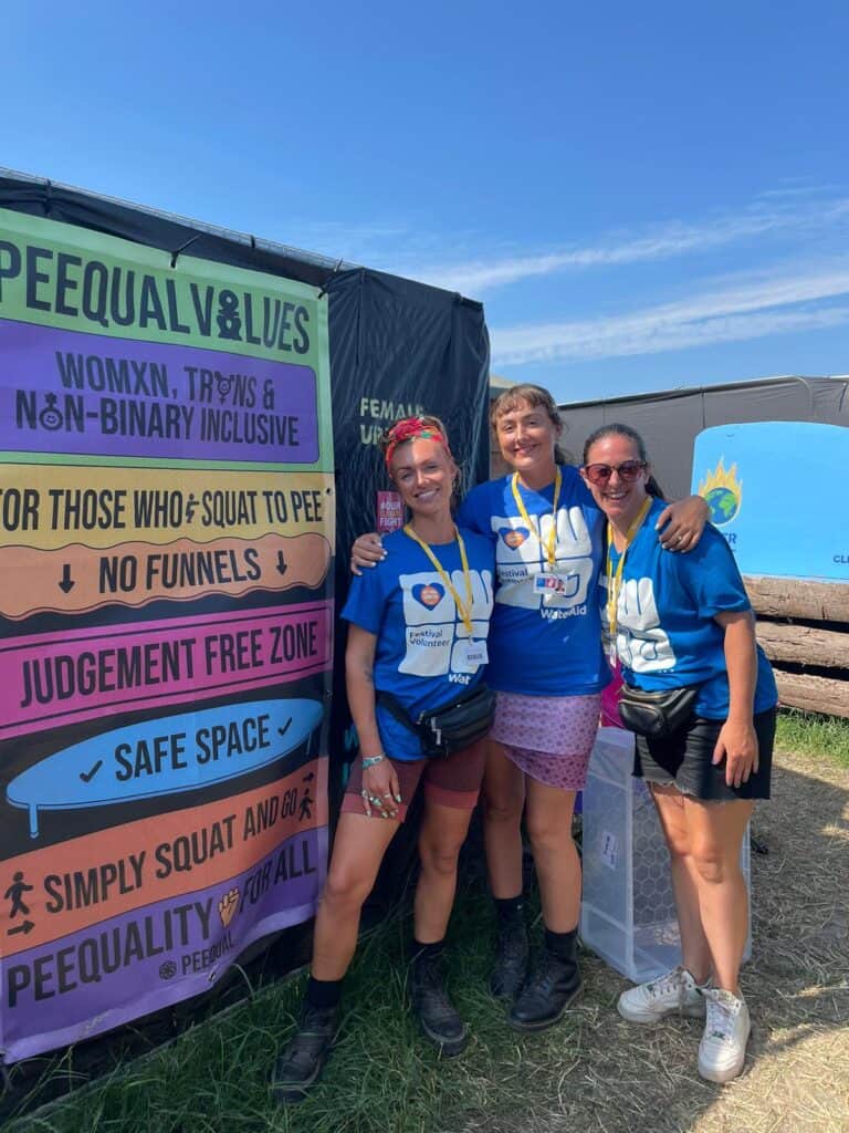Three volunteers wearing festival volunteer and WaterAid T-shirts smiling in front of a colourful banner advocating for inclusive urinals for all genders, promoting a no judgment, safe space for squatting to pee.