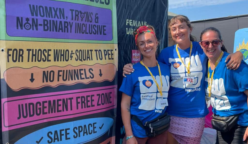 Three volunteers wearing festival volunteer and WaterAid T-shirts smiling in front of a colourful banner advocating for inclusive urinals for all genders, promoting a no judgment, safe space for squatting to pee.