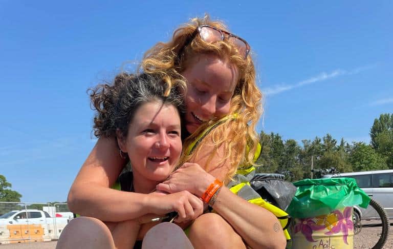Two joyful women embracing in a friendly hug outdoors, one with curly hair in a black tank top and the other wearing a high-visibility vest, both smiling under a clear blue sky.