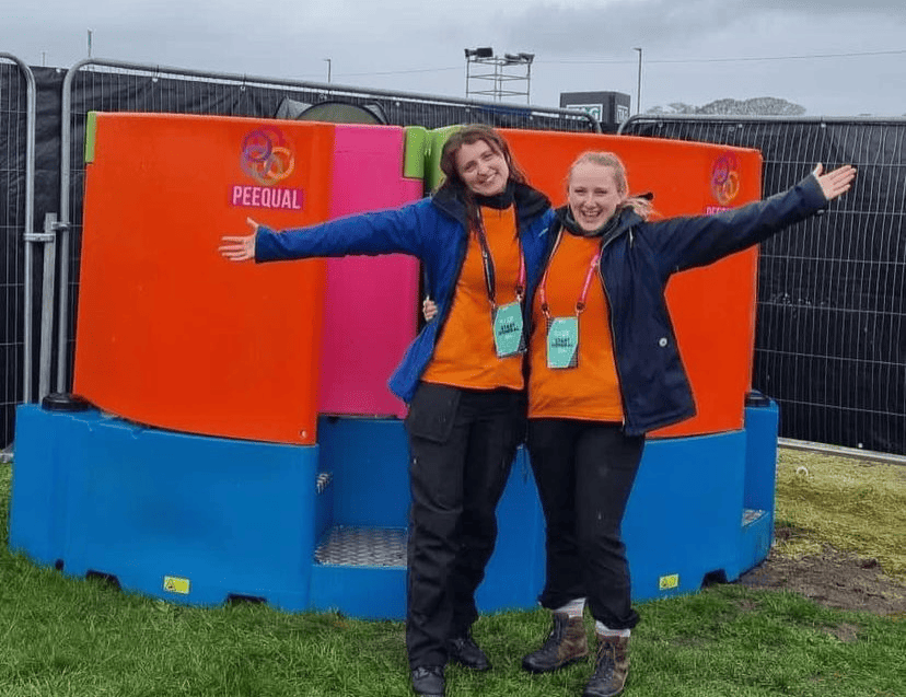 Two smiling individuals in orange crew necks and blue jackets, with arms wide open, standing in front of a multicoloured Peequal urinal setup at an outdoor event.