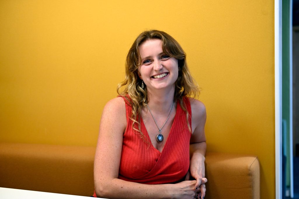 Hazel McShane - a woman with long brown hair, wearing a red top and chain necklace against a yellow backdrop.