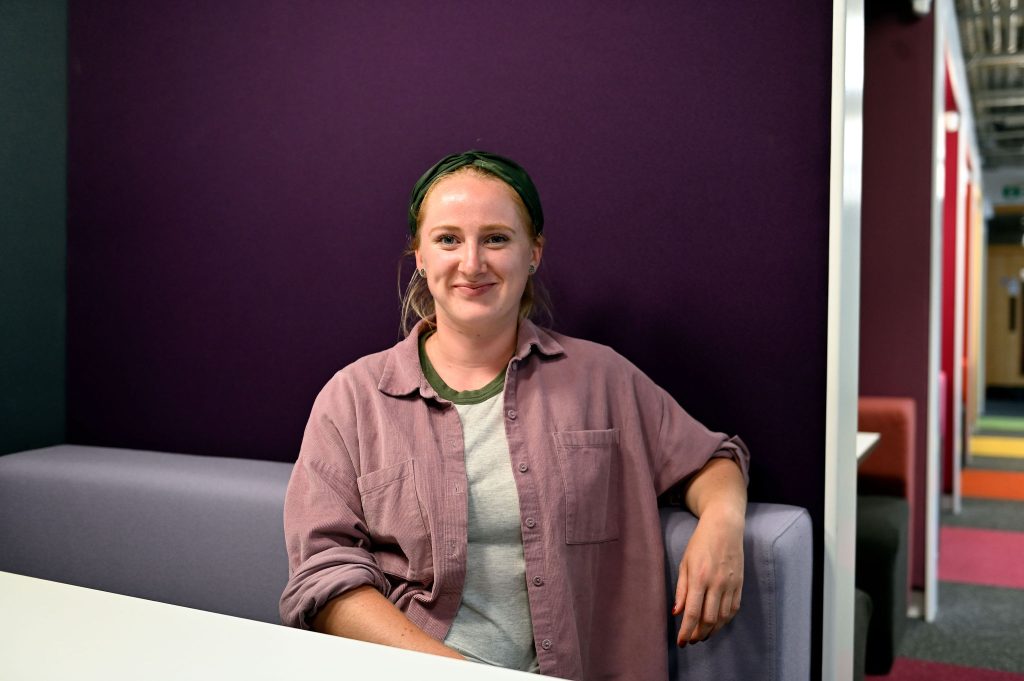 A woman wearing a green headscarf and light purple top against a dark purple backdrop.