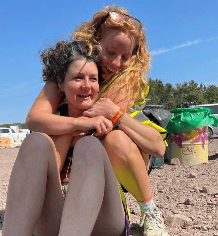 Two joyful women embracing in a friendly hug outdoors, one with curly hair in a black tank top and the other wearing a high-visibility vest, both smiling under a clear blue sky.