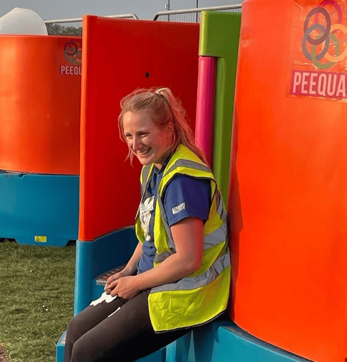 A smiling woman with blonde hair, wearing a high-visibility vest, sits on the step of a vibrant, multicoloured Peequal urinal.