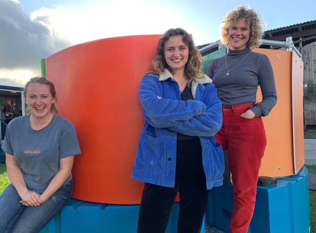 Three smiling women standing confidently in front of a colourful Peequal urinal setup, with one seated on the blue base, another crossing arms wearing a blue jacket, and the third with hands on hips in red trousers.