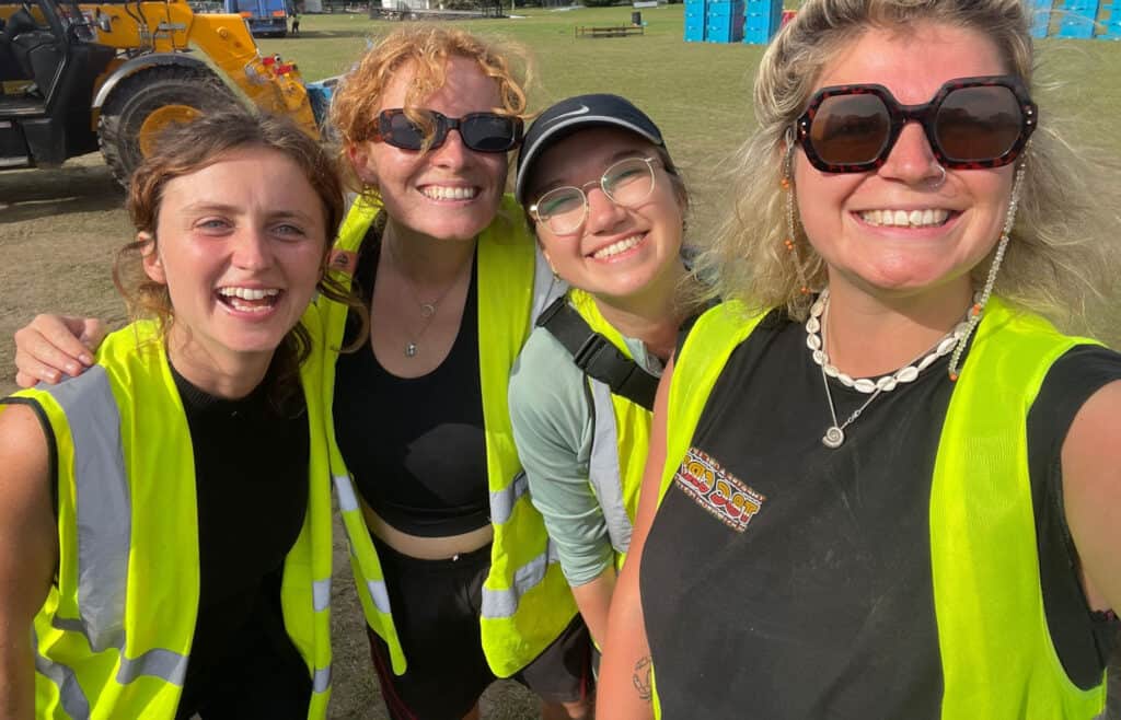 Four cheerful women in high-visibility vests taking a group selfie, with a construction vehicle and portable toilets in the background at an outdoor event.