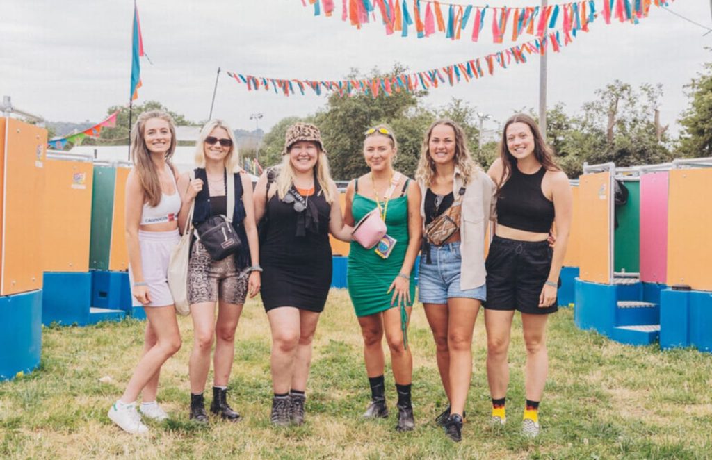 A group of festival-goers in colourful clothing outside a row of Peequal urinals, posing under colourful festival bunting.