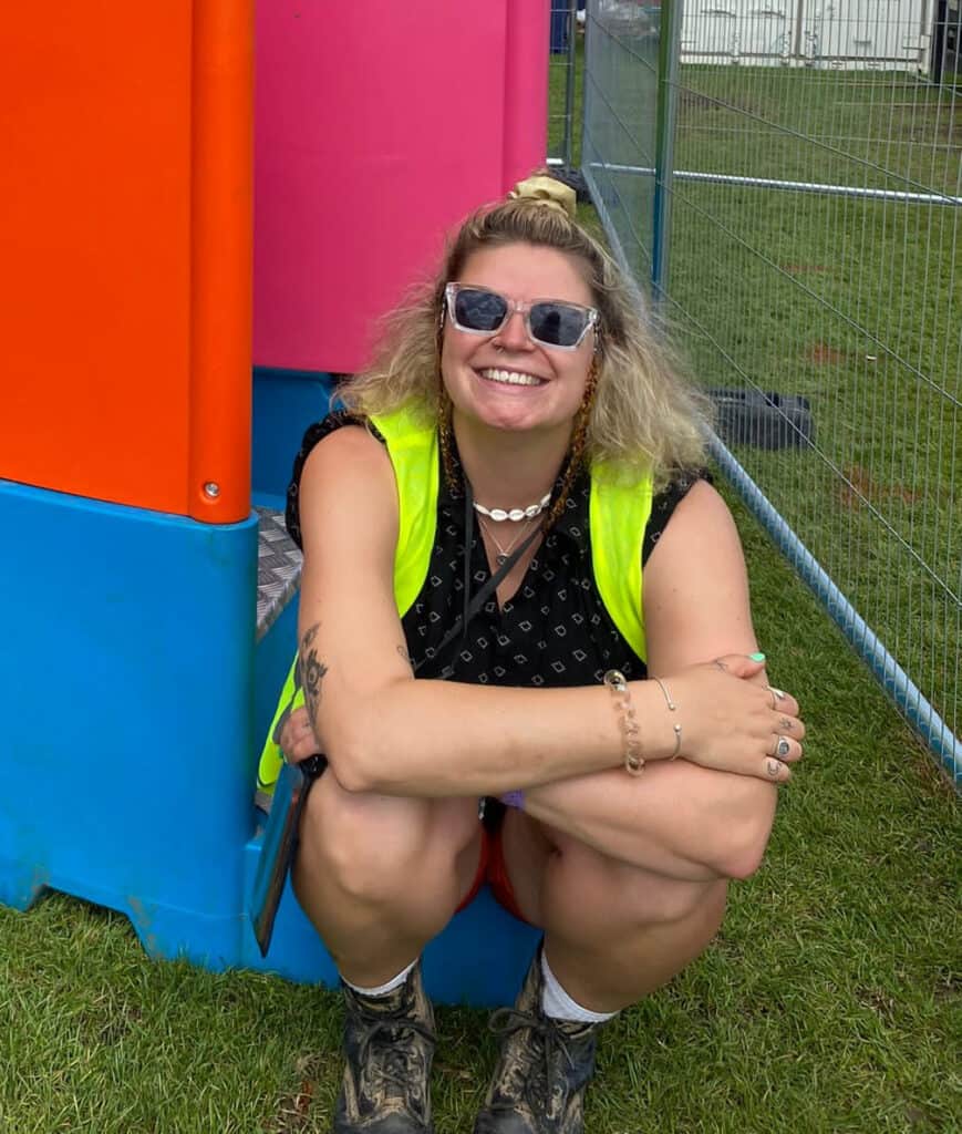 A happy blonde haired woman sitting in front of a colourful Peequal urinal setup, wearing a high-visibility vest and sunglasses at an outdoor festival location.