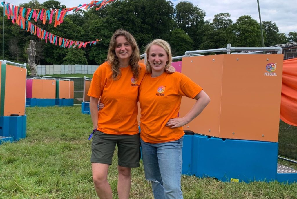 Two women in orange t-shirts standing outside a row of Peequal urinals.