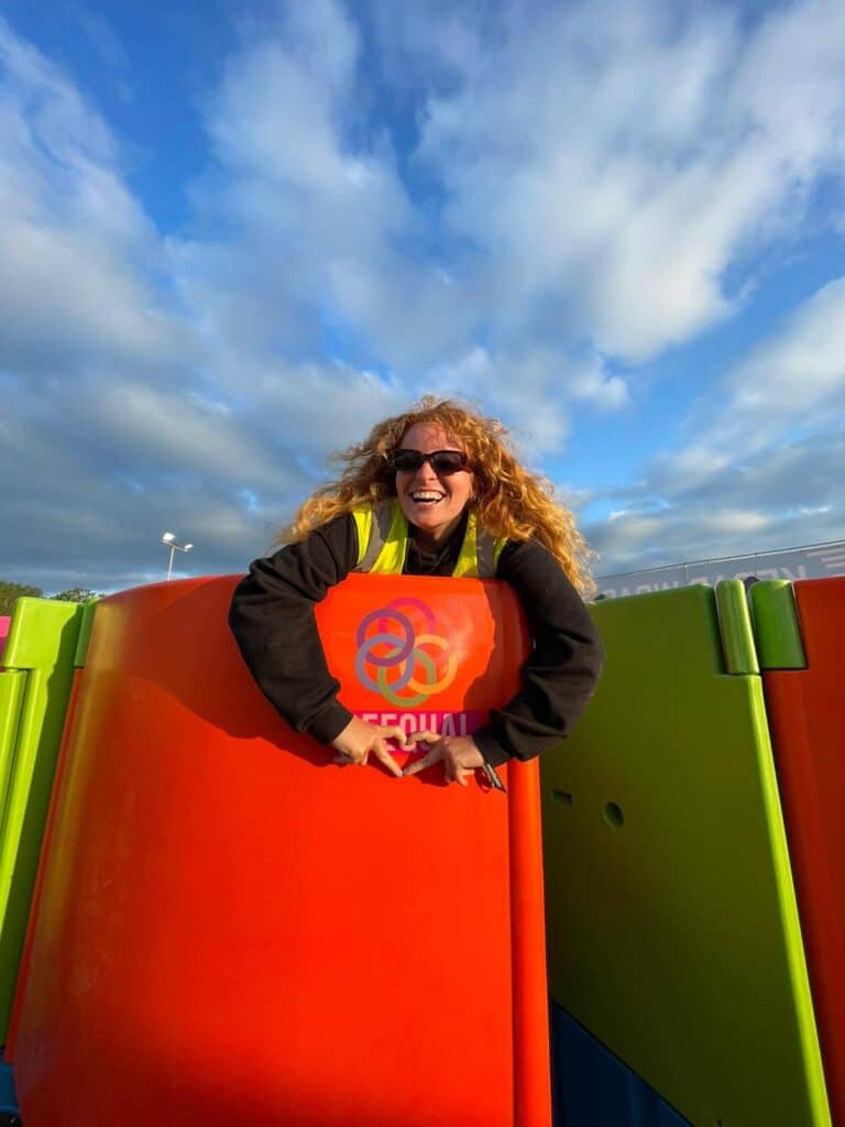 A joyful woman in a high-visibility vest leans over the top of a vibrant orange Peequal urinal, with a clear blue sky in the background.