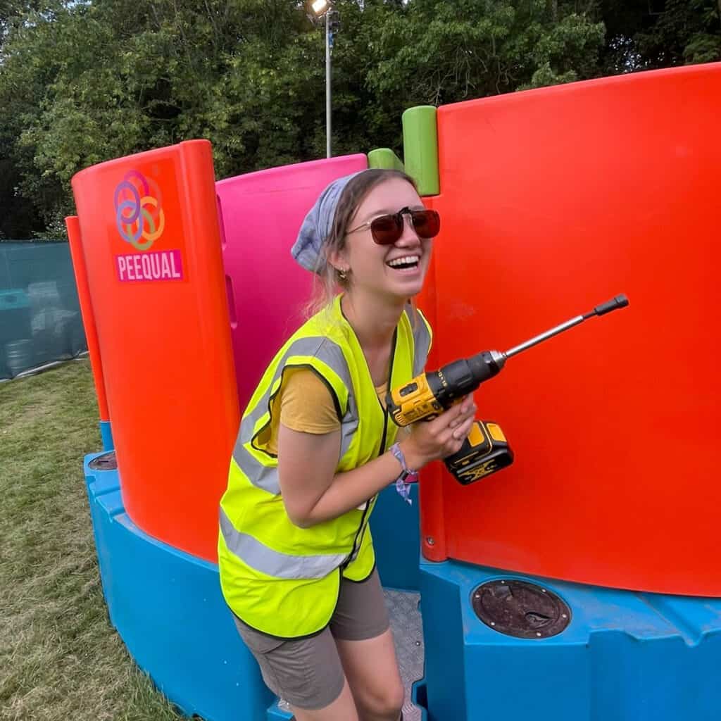 A woman in a high-visibility vest and sunglasses is laughing while holding a drill, standing next to a brightly coloured Peequal urinal partition outdoors.