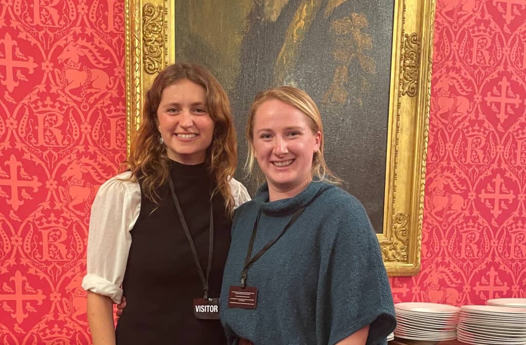 Two smiling women with visitor badges standing indoors in front of a decorative pink wallpaper with a classic pattern.