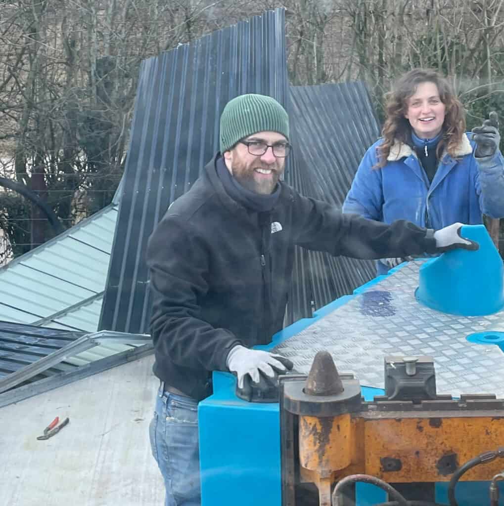 A man and a woman, both smiling and wearing warm clothing and gloves, work together on assembling a blue Peequal urinal in an outdoor setting with metal structures in the background.