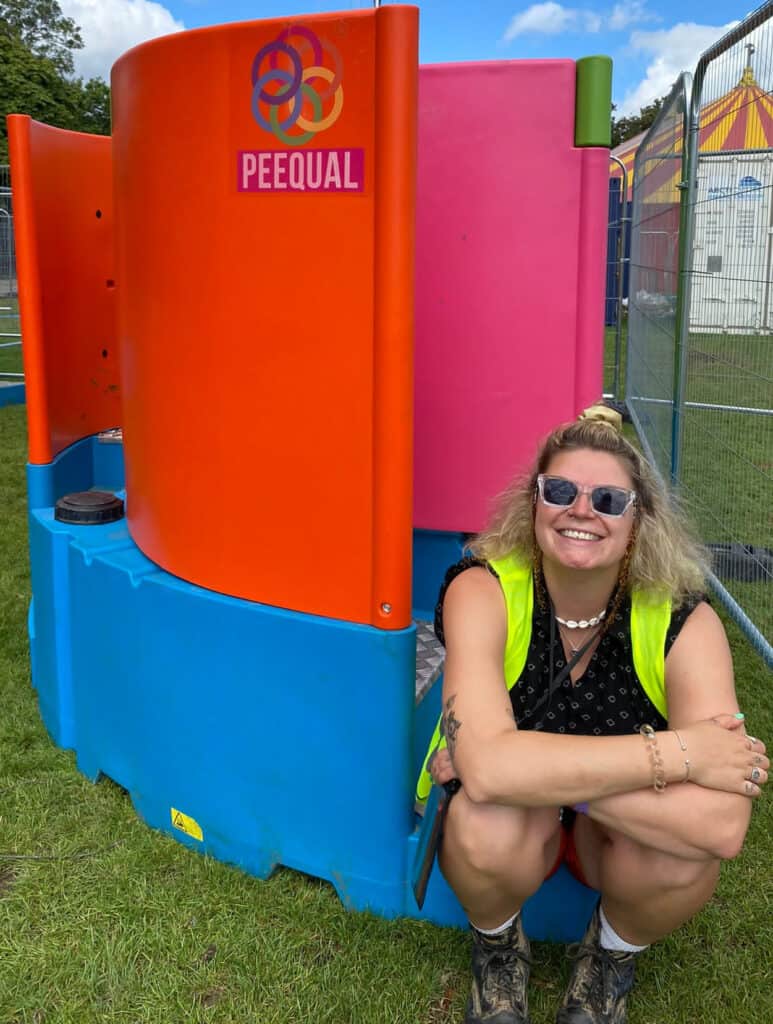 A happy blonde haired woman sitting in front of a colourful Peequal urinal setup, wearing a high-visibility vest and sunglasses at an outdoor festival location.
